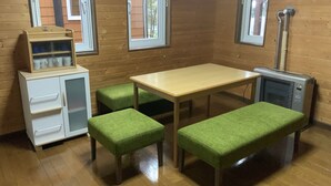 A room with a wooden floor, a table with green upholstered benches, a white cabinet with drawers, and a window with a view of trees. - Ninjaart Furano Cottage (Furano)