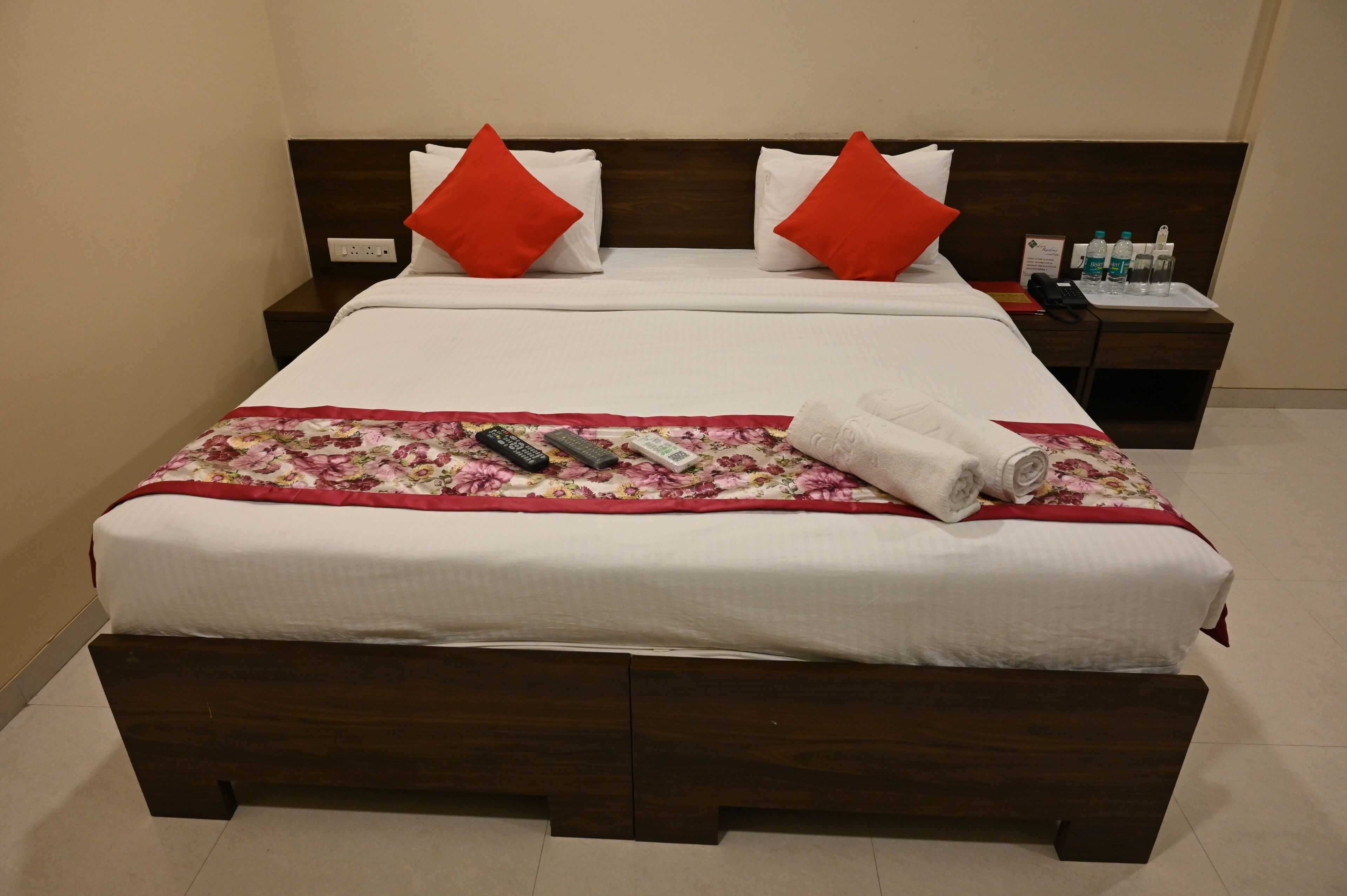 A neatly made bed with a floral bedspread, two red pillows, and a rolled towel on the side.