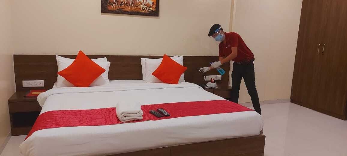 Una cama bien hecha con un edredón rojo y dos almohadas naranjas, una mesita de noche con un libro y mandos a distancia, y una persona limpiando el suelo.