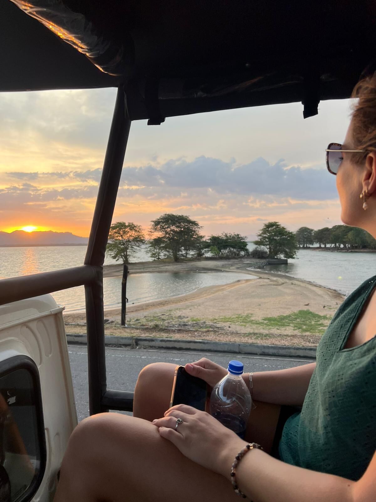 A person seated inside a vehicle, holding a smartphone and a water bottle, with a view of a sunset over a body of water and trees.