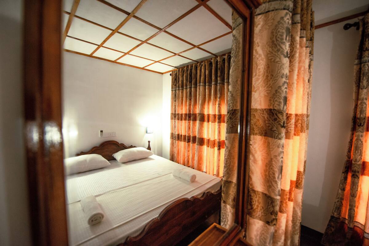 A neatly made bed with a wooden headboard, a bedside table with a lamp, and patterned curtains.