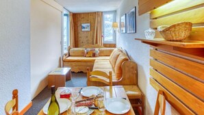 Une pièce chaleureuse avec un canapé en bois, une table à manger dressée avec des assiettes et une bouteille, et une fenêtre avec des rideaux.