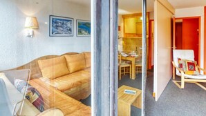 Un salon confortable avec un canapé, une petite table et un fauteuil. On aperçoit un coin repas avec une table et des chaises à travers une porte ouverte.