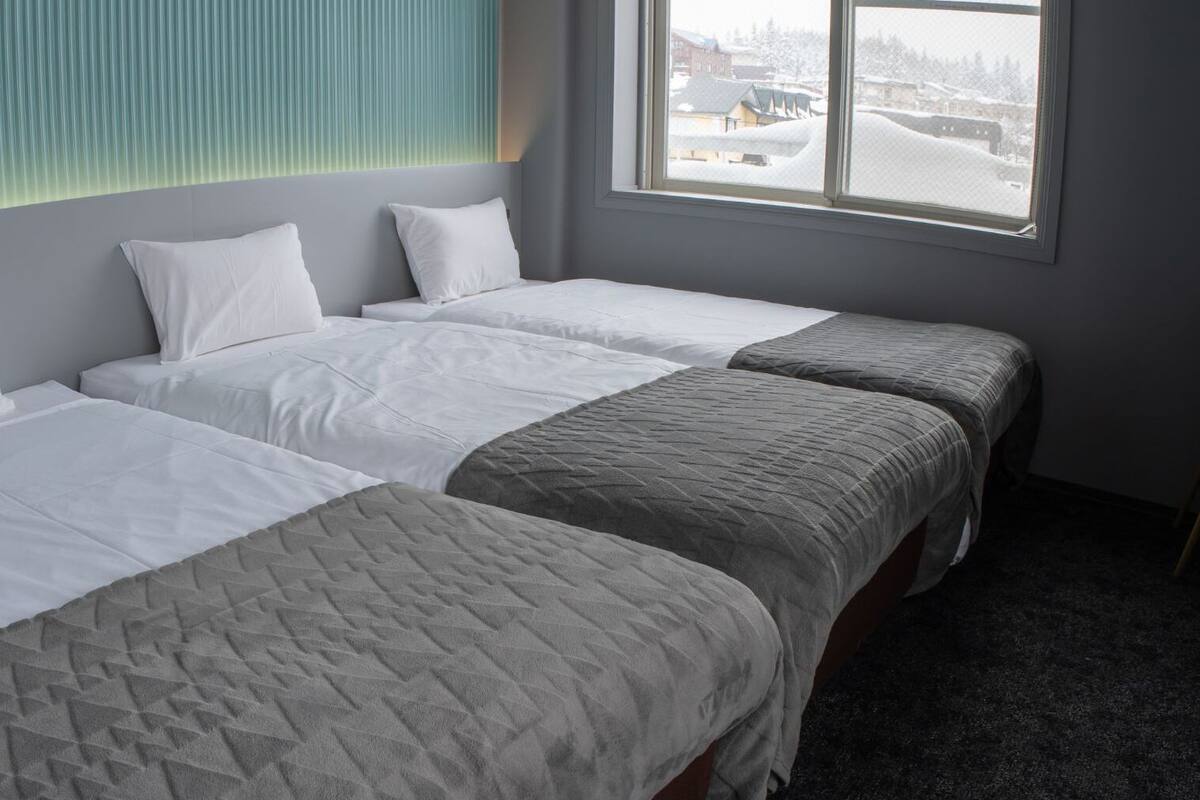 Ein Hotelzimmer mit drei Betten, einem Fenster mit Blick auf eine verschneite Landschaft und einer Wand mit strukturierter Verkleidung.