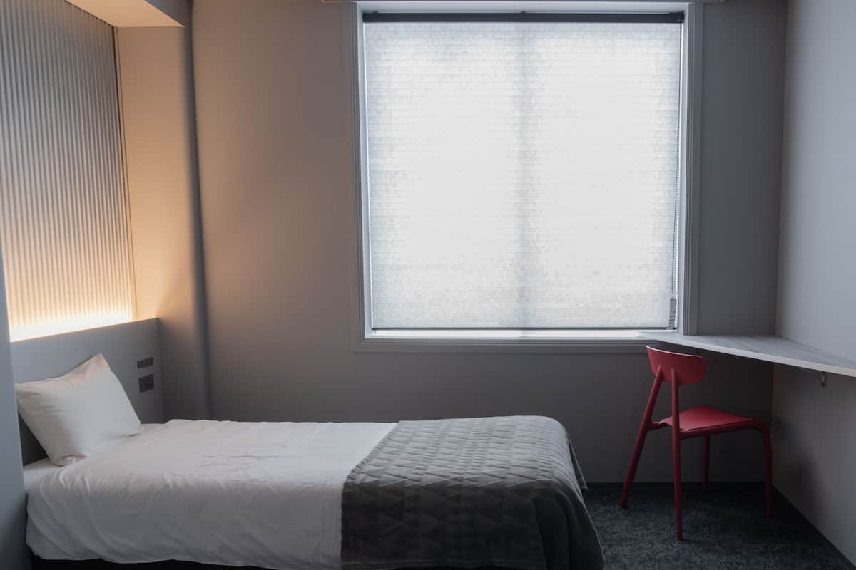 Ein Schlafzimmer mit einem Bett, einem Fenster mit Jalousien, einem roten Sessel und einem kleinen Schreibtisch.