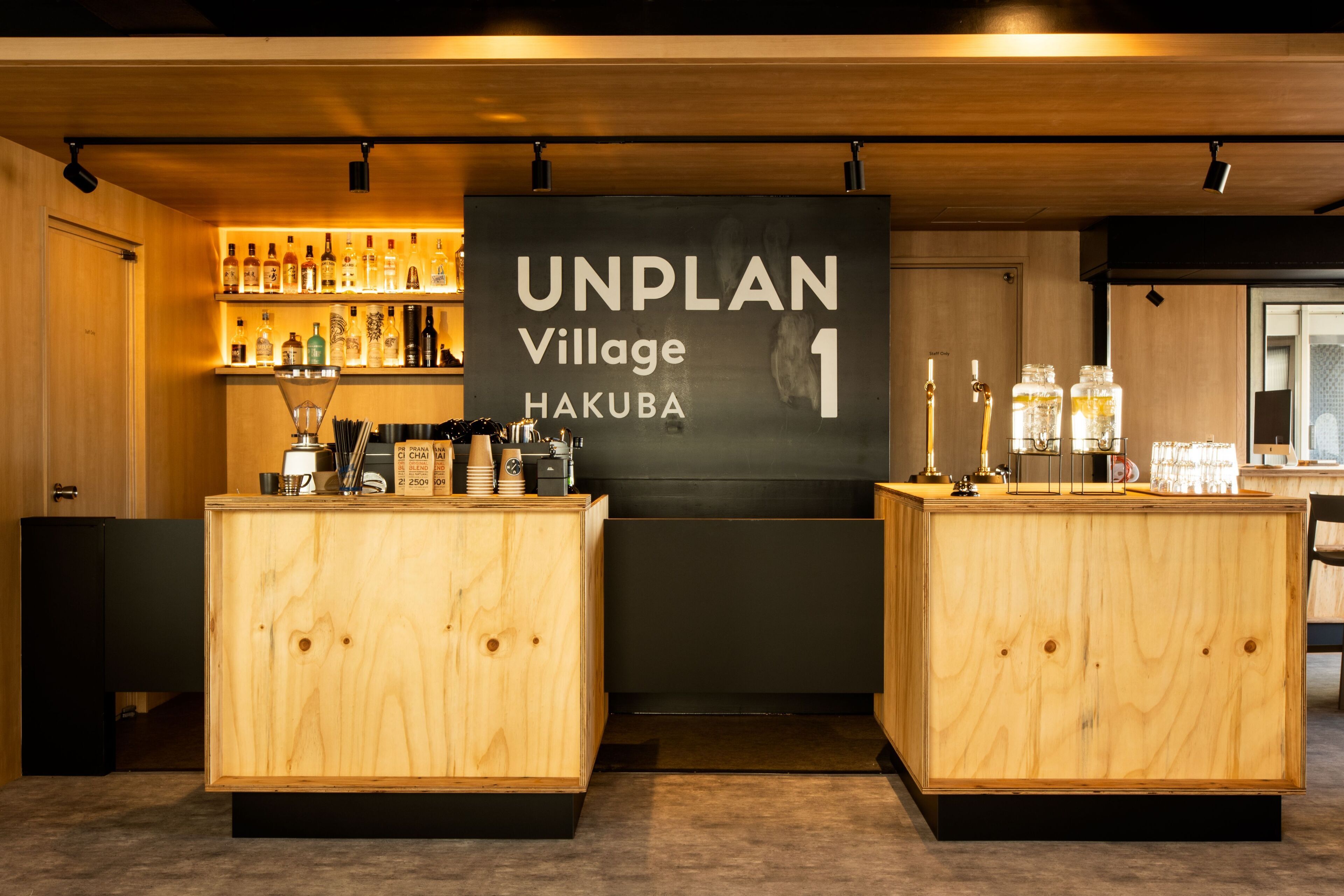 A bar area with wooden cabinets, shelves of liquor bottles, and a sign reading "UNPLAN Village Hakuba".