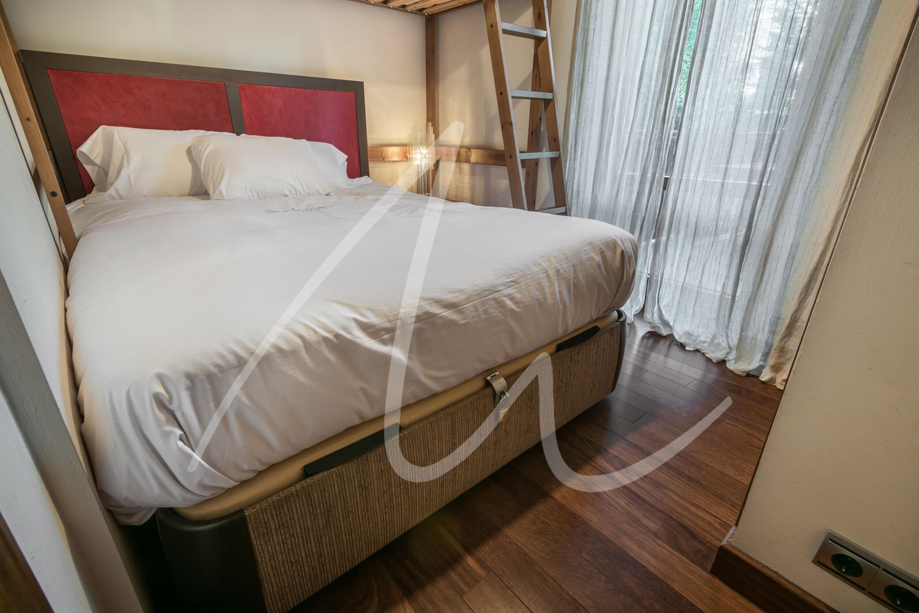 A bed with a red headboard, a wooden ladder, and a window with sheer curtains.