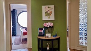 A hallway with a green wall, a round mirror, a small table with a vase of flowers, and a door leading to a room with a bed and curtains.