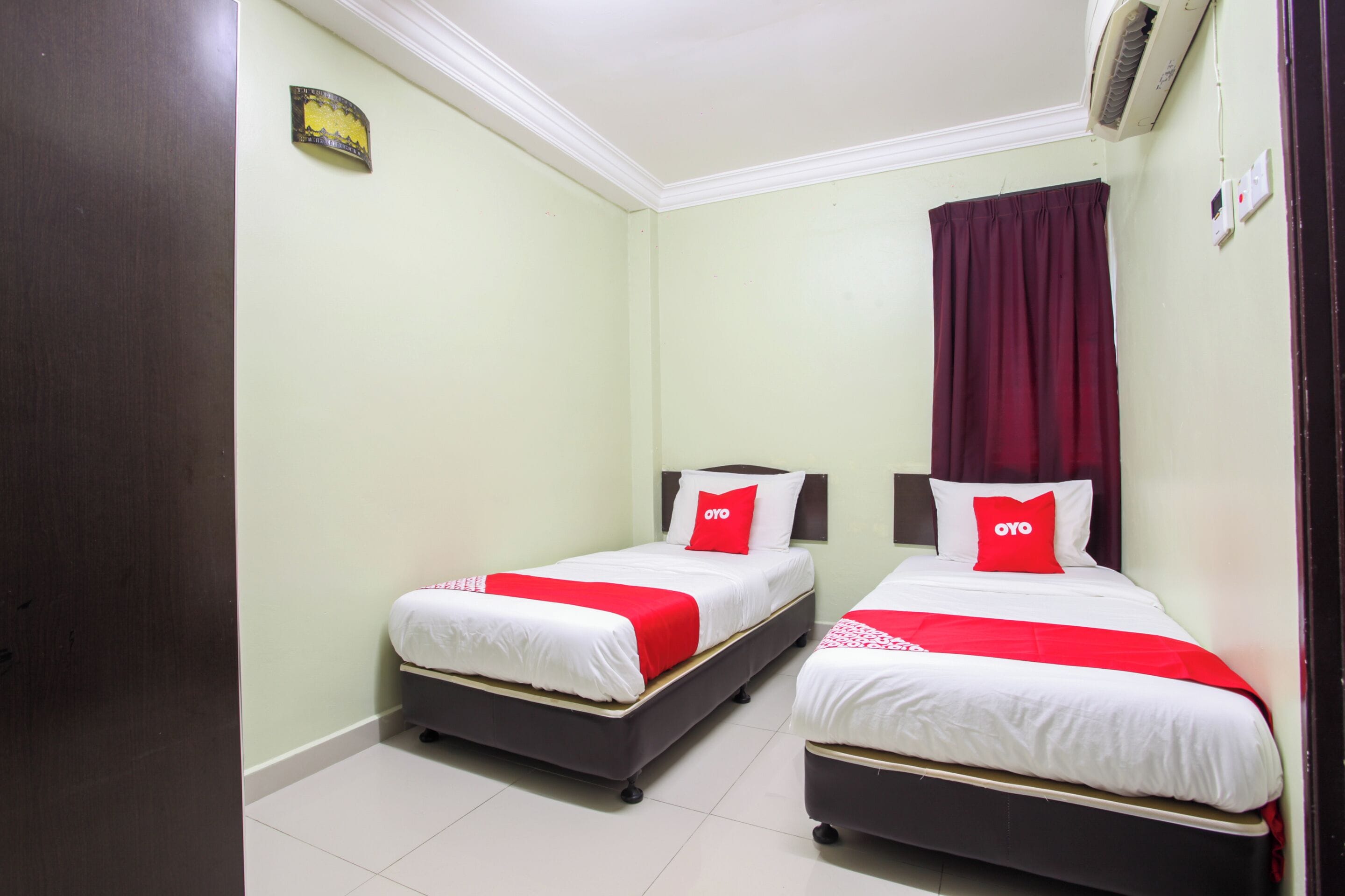 Habitación de hotel con dos camas, cada una con ropa de cama blanca y detalles en rojo, una cortina roja y un aire acondicionado de pared.