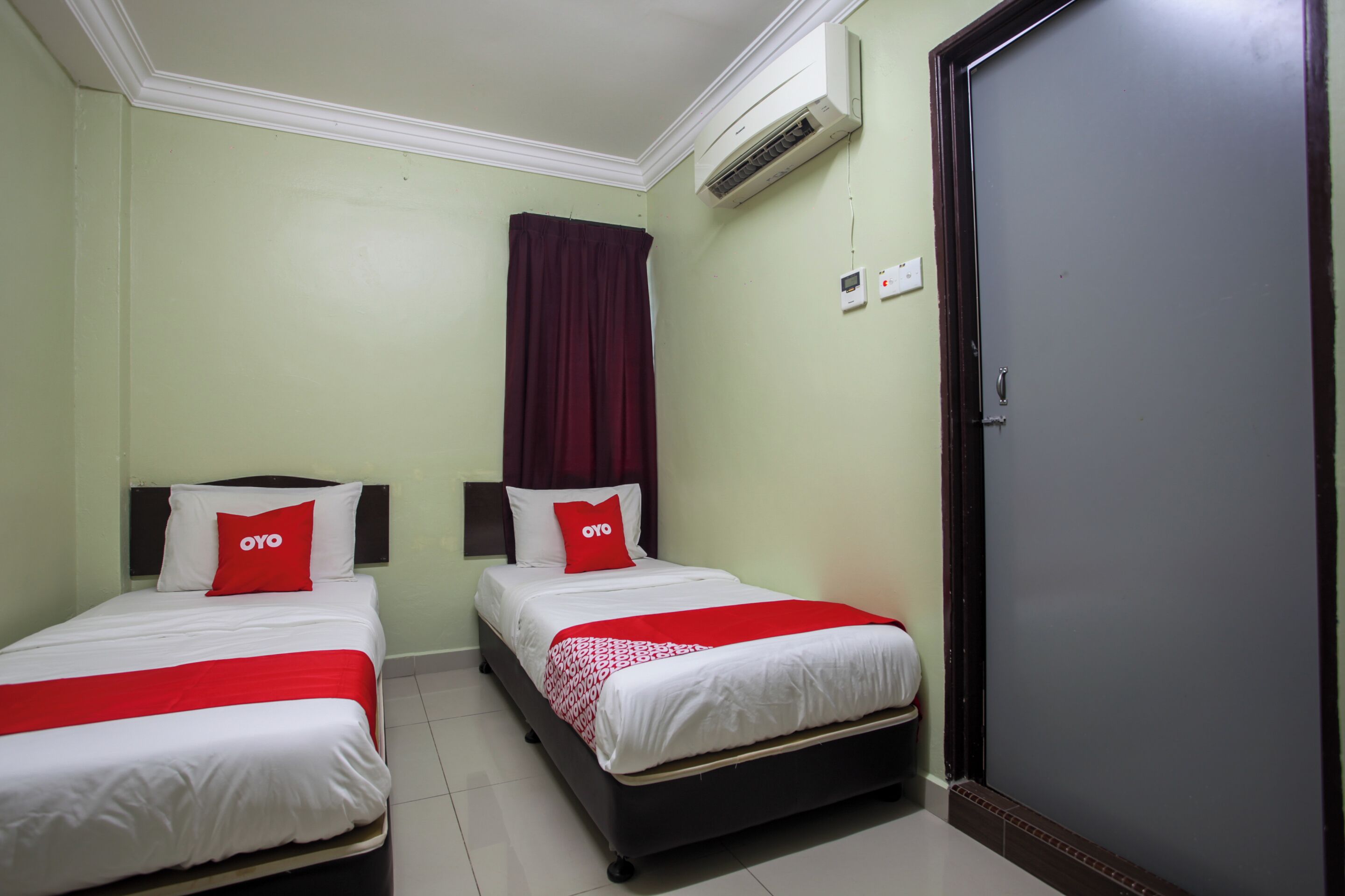 A hotel room with two beds, each with white bedding and red accents, a red curtain, and an air conditioning unit on the wall.