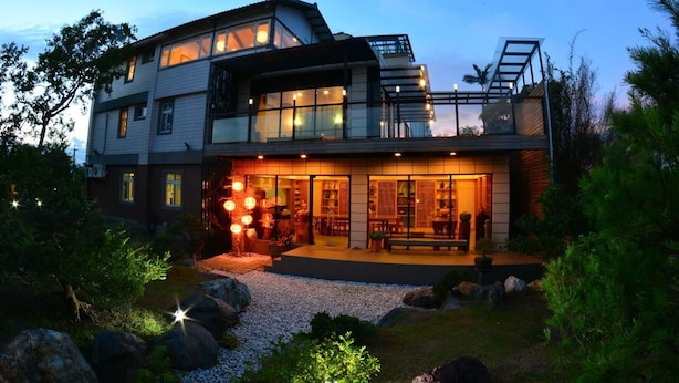 Une maison moderne à deux étages, dotée d’une grande façade vitrée, d’un espace extérieur aménagé et couvert, et d’un jardin bien entretenu avec des rochers et des plantes.