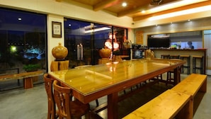 Un espace repas moderne avec une grande table en bois et des bancs, un coin cuisine équipé d’un téléviseur, et une vue sur le ciel nocturne grâce à de grandes fenêtres.