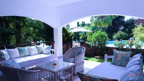 Un espacio exterior soleado con mobiliario de mimbre, una mesa de vidrio y vistas a una piscina y abundante vegetación.
