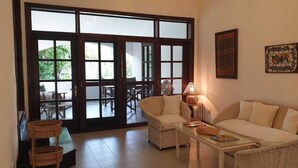 Sala de estar con sofá, sillones, una mesa de centro y una lámpara. Hay una puerta corrediza de vidrio que da a un espacio exterior con una mesa y sillas.