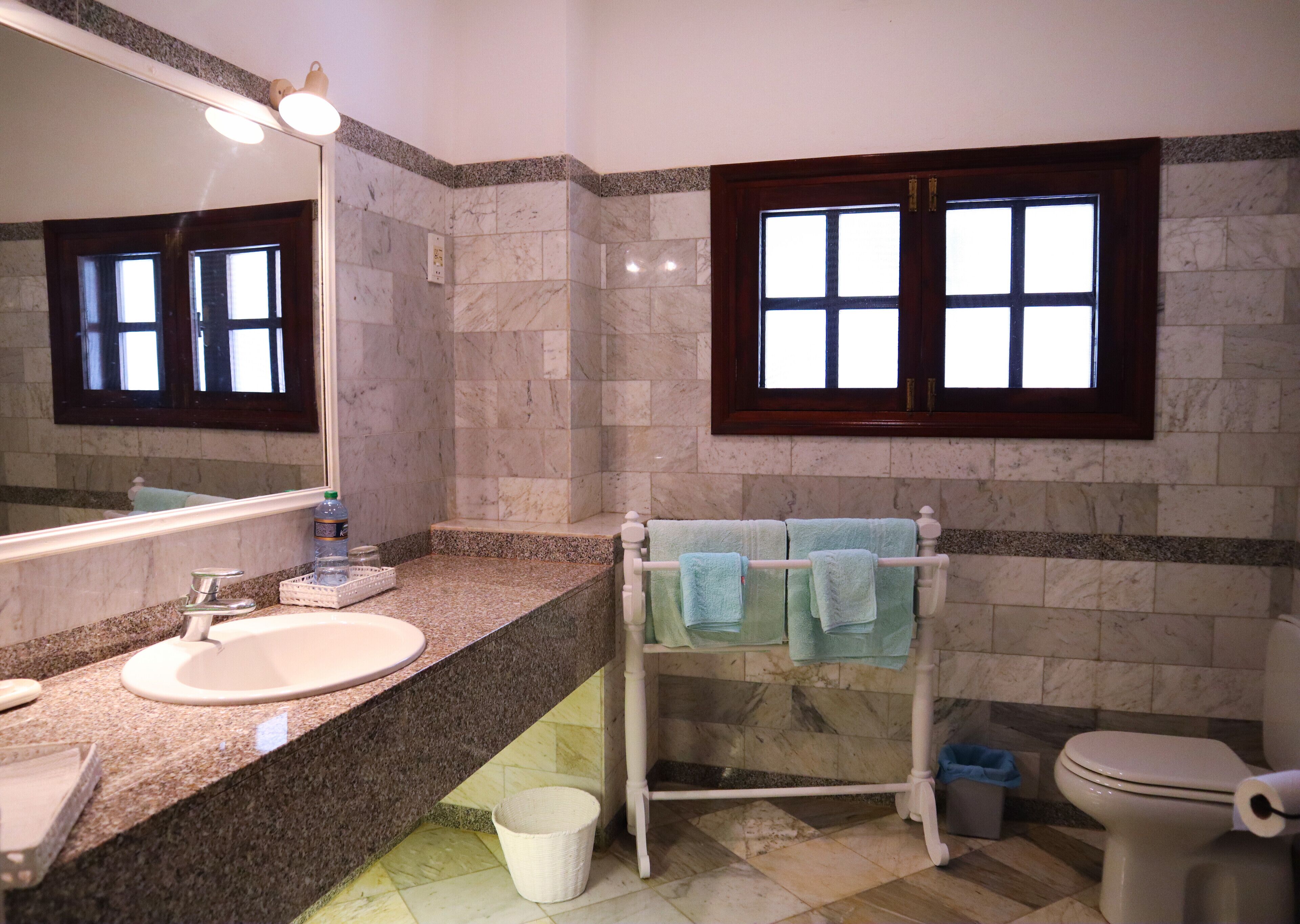A bathroom with a granite countertop sink, a mirror, a towel rack with towels, a toilet, and a window with a wooden frame.