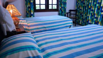 Una habitación con dos camas, una mesita de noche con lámpara, una silla y un ventanal con cortinas.
