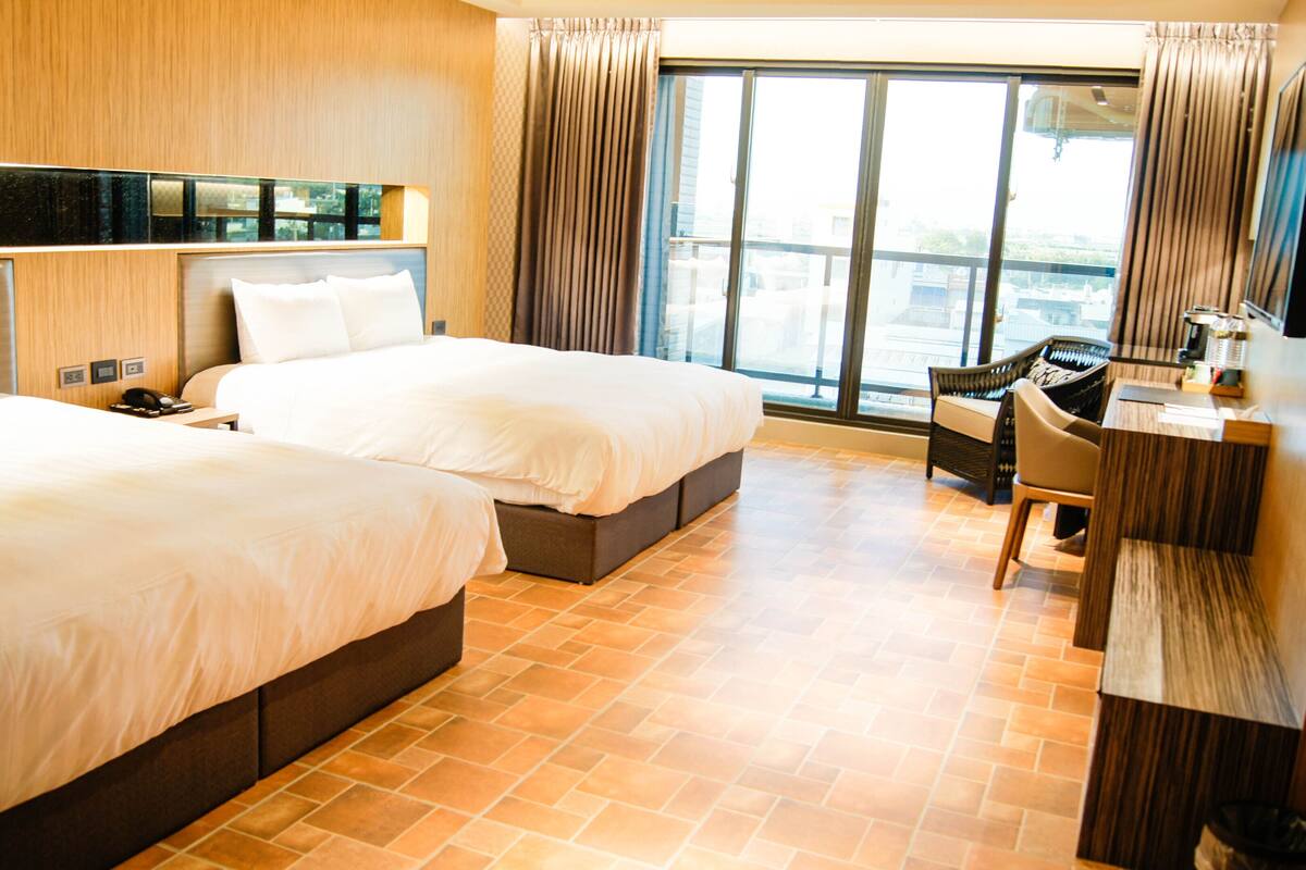 A hotel room with two beds, a desk, a chair, and a large window with curtains.
