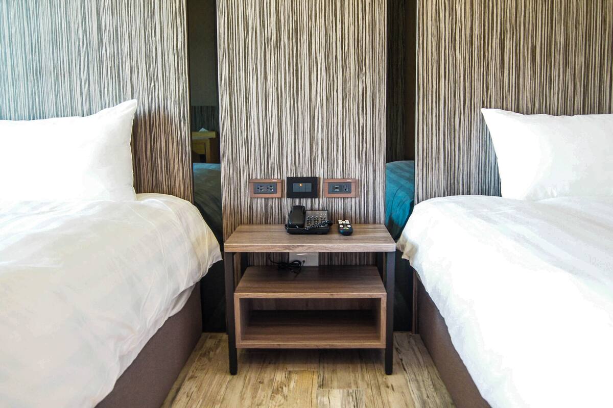 A hotel room with two beds, a wooden nightstand with a telephone and remote control, and a wall with vertical lines.