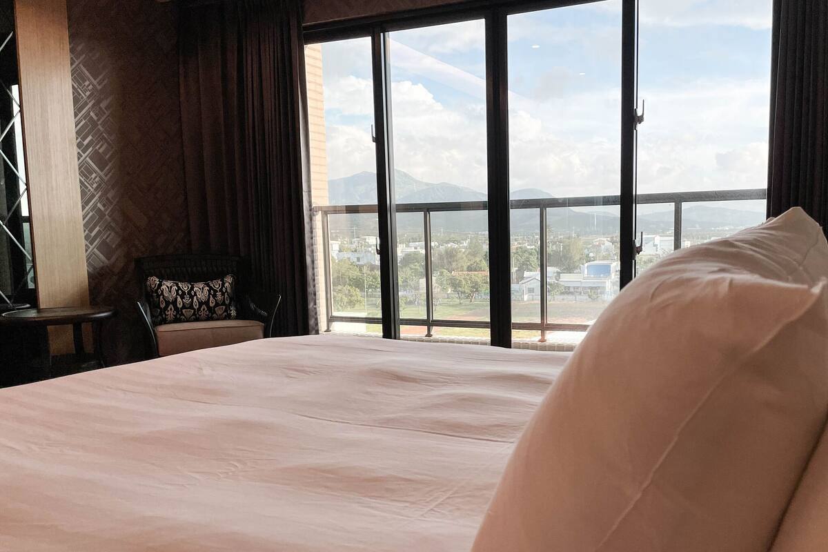 A hotel room with a large bed, a view of the city through floor-to-ceiling windows, and a balcony with a railing.