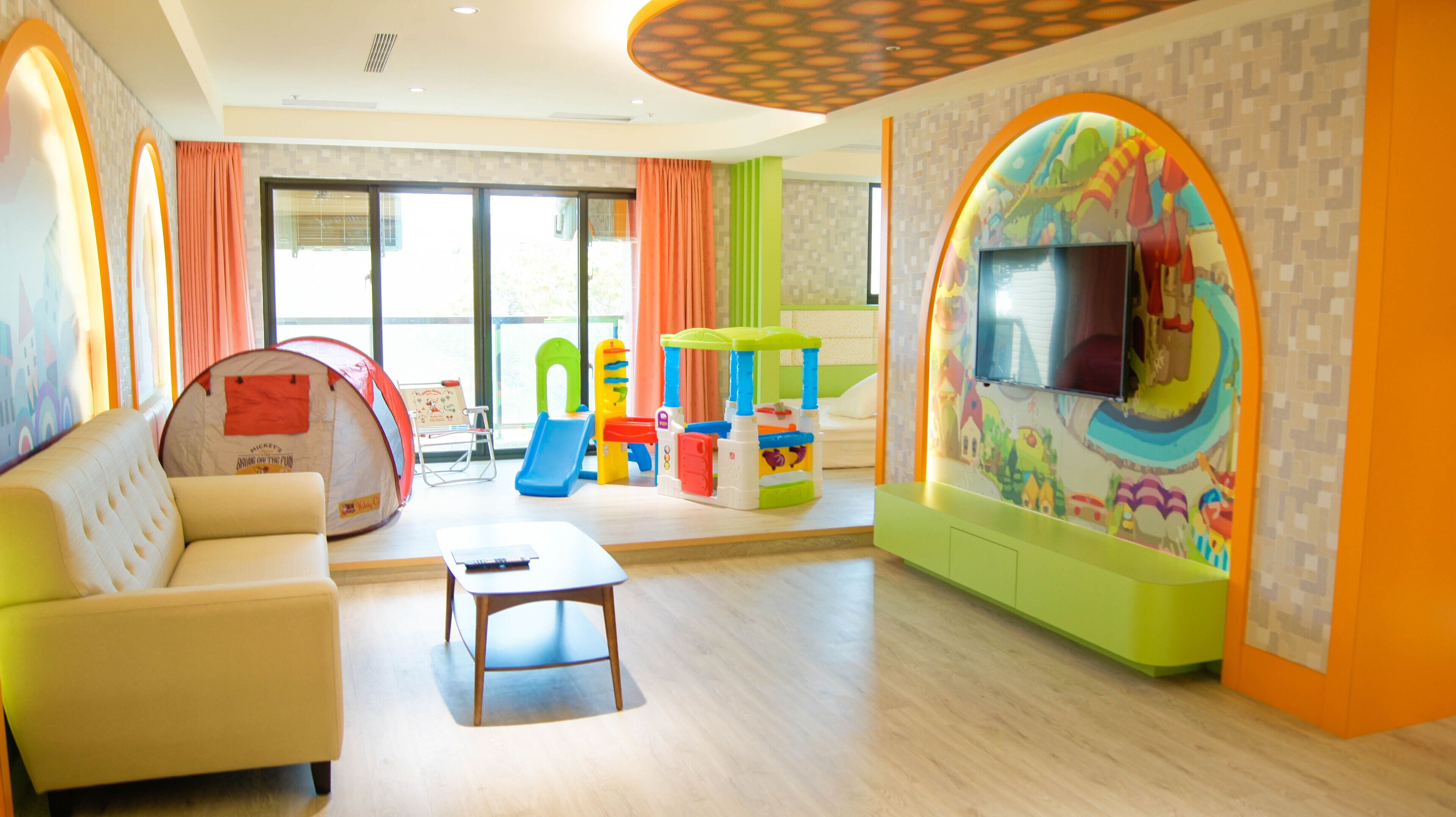 A spacious living room with a sofa, coffee table, and colorful play structures. A television is mounted on a wall with a playful mural.