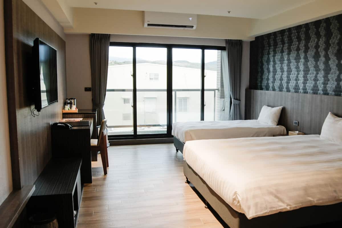 A hotel room with two beds, a desk, a chair, a flat-screen TV, and a large window offering a view of the city.