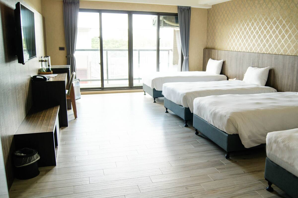 A hotel room with three beds, a TV, and a large window with curtains.