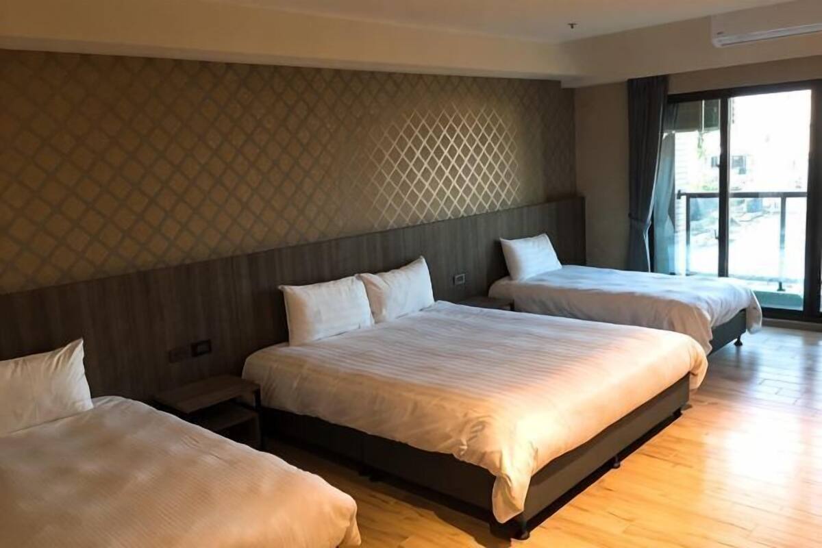 A hotel room with three beds, wooden flooring, a patterned wall, and a large window.