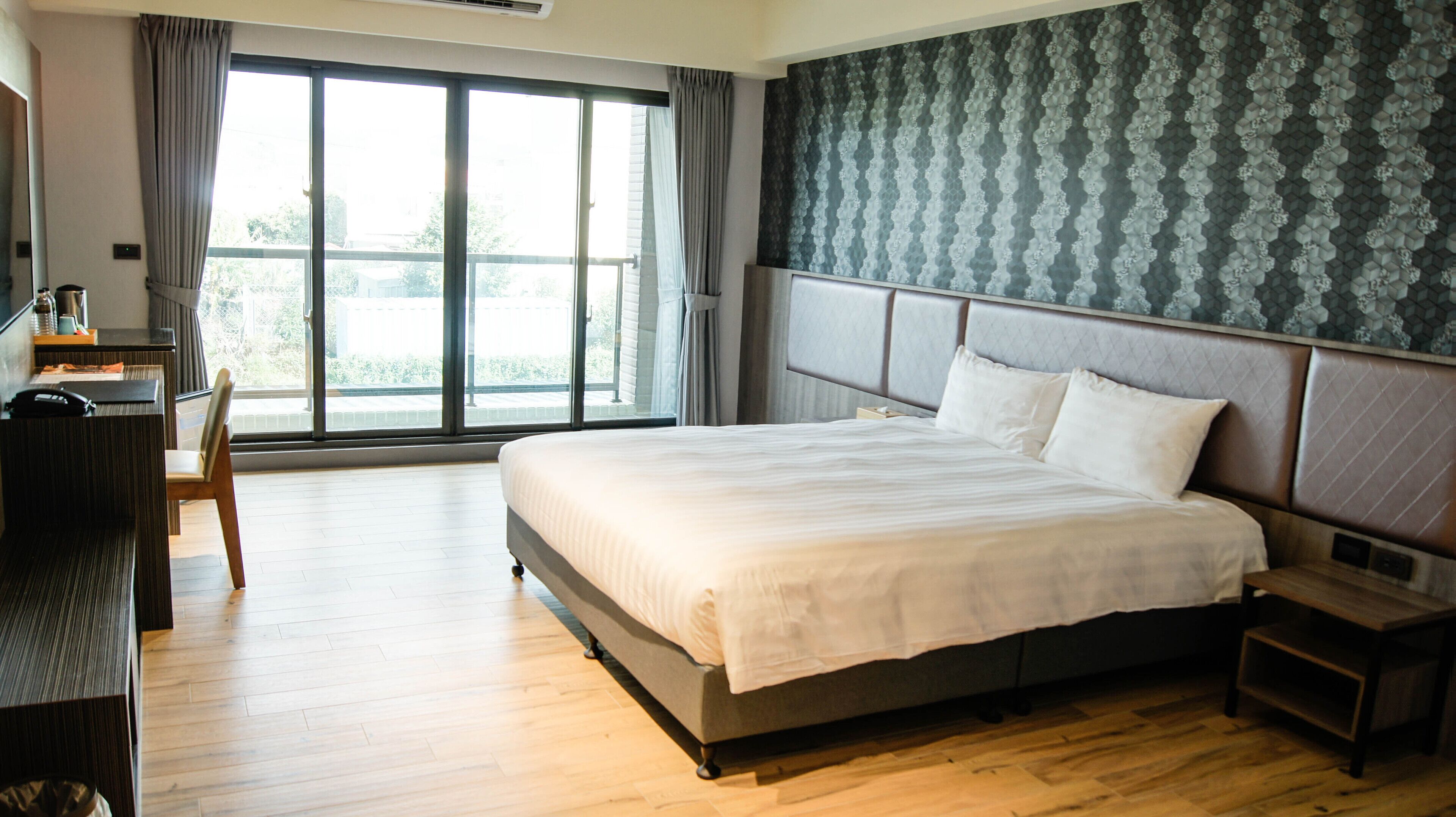 A hotel room with a large bed, a desk, and a chair. There is a window with curtains, and a patterned wallpaper on the wall.