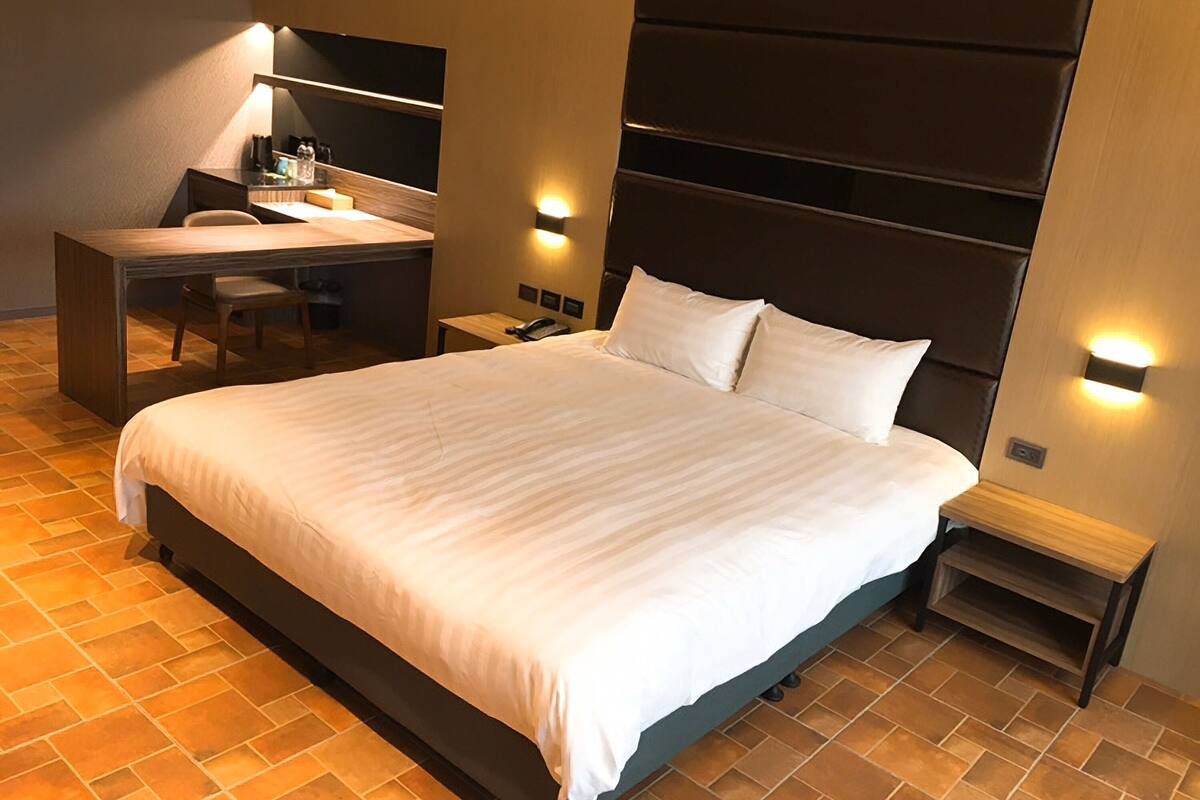 A hotel room with a large bed, a desk with a chair, a mirror, and two bedside tables with lamps.