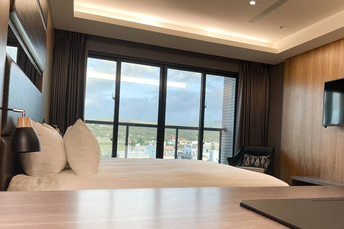 A hotel room with a large bed, a TV mounted on the wall, and a view of a cityscape through large windows.
