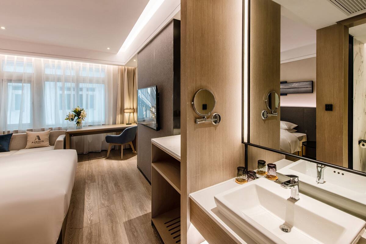 Une chambre d’hôtel comprenant un lit, un bureau, une chaise, un grand miroir, une salle de bain avec un lavabo et des toilettes, et une fenêtre avec des rideaux.