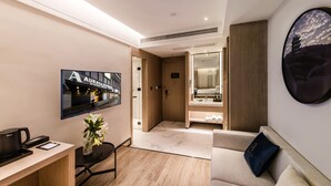 A modern hotel room with a flat-screen TV, a desk with a coffee machine, a sofa with a pillow, and a large mirror reflecting the room. - Zhangjiagang Suning Auraya Hotel (Suzhou)
