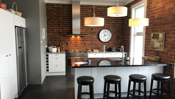A modern kitchen with a brick wall, stainless steel appliances, a central island with stools, pendant lighting, and a clock on the wall. - Top floor 3 bed apartment in the iconic Tiger Tea Building ( FREE Netflix) (Dunedin)