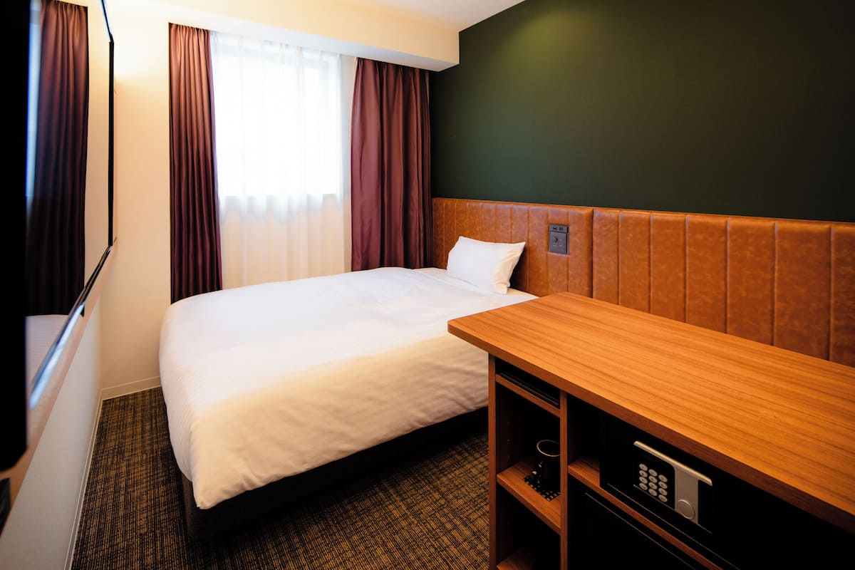 A hotel room with a large bed, a wooden desk, a built-in safe, and a window with curtains.