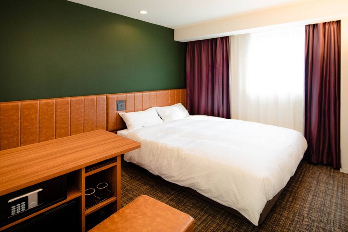 A hotel room with a bed, a wooden desk, a TV, and a window with curtains.