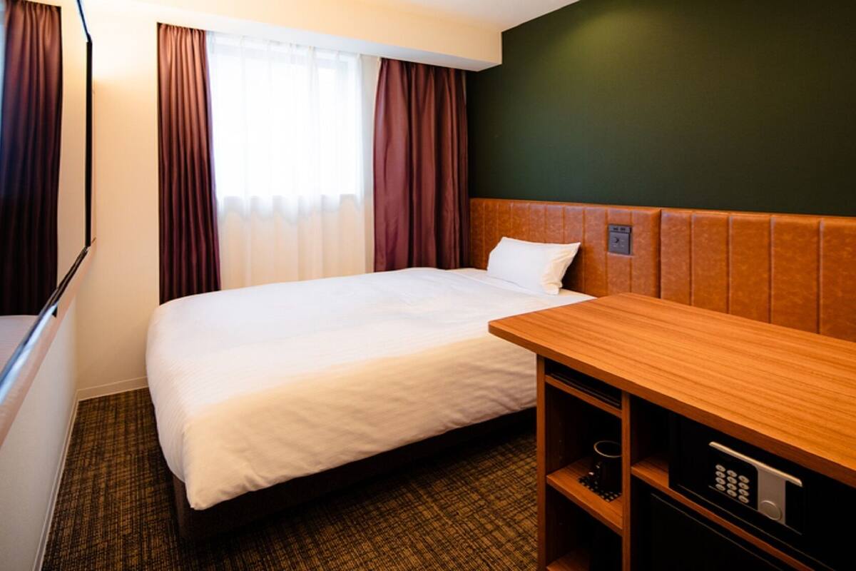 A hotel room with a bed, a wooden desk, a small shelf, and a control panel.
