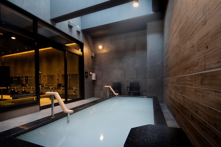 A modern indoor pool with clear glass walls, wooden paneling on one side, and a seating area with chairs.