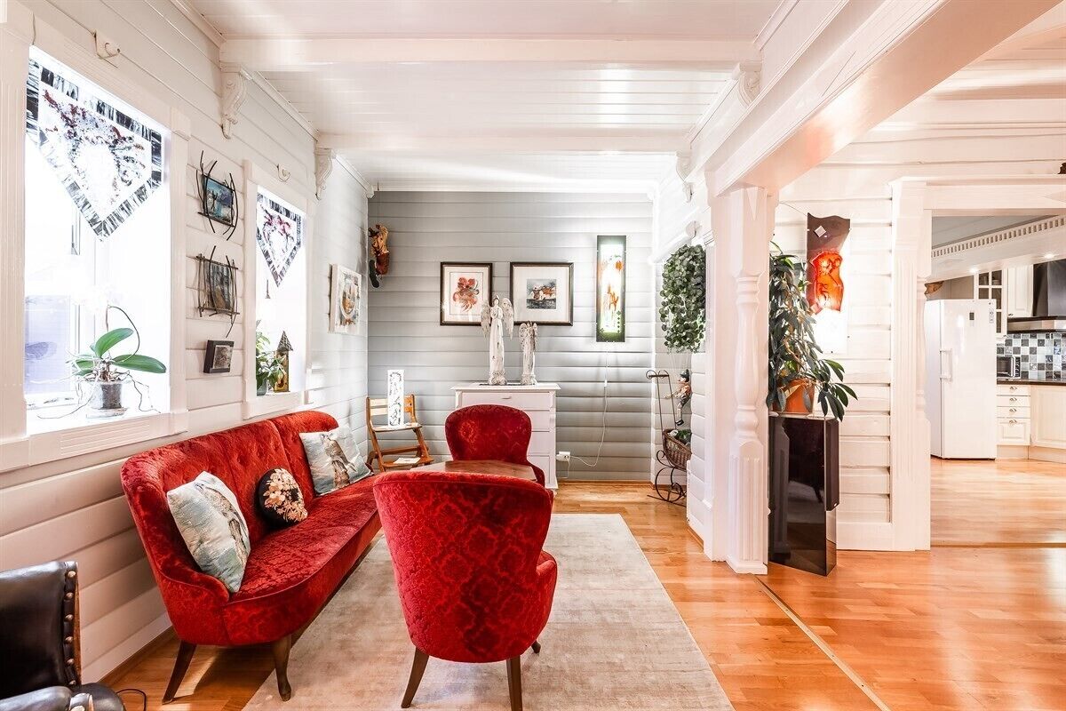 A spacious living room with a red sofa and armchair, a white fireplace, and a kitchen visible through an open doorway.