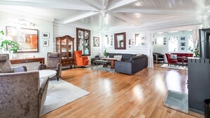Een ruime woonkamer met houten vloeren, een hoog plafond met zichtbare balken en een open haard. Er zijn meerdere zithoeken met banken en fauteuils.