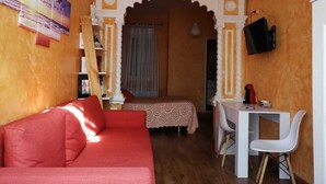 A cozy room with a red sofa, a small desk with a chair, a bed with a patterned bedspread, and a TV mounted on the wall. - Hospedaje Menendez Pelayo (Santander)