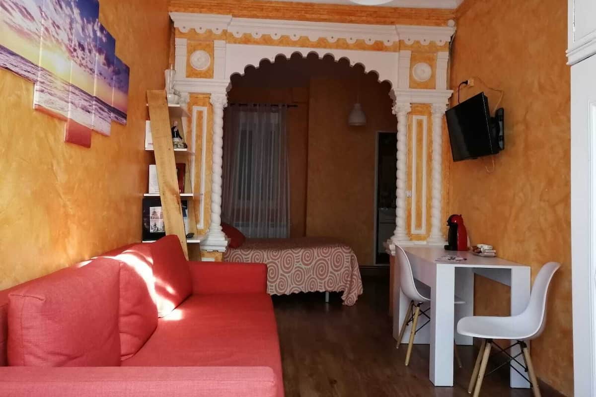 Ein gemütliches Zimmer mit einem roten Sofa, einem kleinen Schreibtisch mit Stuhl, einem Bett mit gemustertem Überwurf und einem an der Wand befestigten Fernseher.