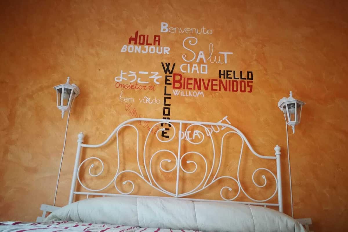 Ein Bett mit einem weißen Metall-Kopfteil und einer Wand mit mehrsprachigen Willkommensschildern.