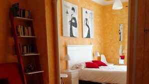 A bedroom with a wooden floor, a red chair, a white bed, a nightstand, a bookshelf, and two wall art pieces. - Hospedaje Menendez Pelayo (Santander)