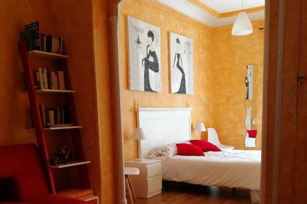 Ein Schlafzimmer mit Holzboden, einem roten Sessel, einem weißen Bett, einem Nachttisch, einem Bücherregal und zwei Wandbildern.