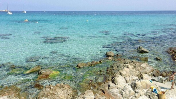 Uma costa rochosa com águas turquesa cristalinas, barcos ao longe e uma pessoa de biquíni.
