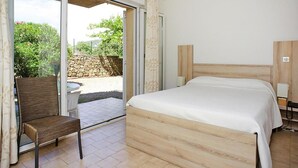 Quarto com cama de madeira, uma cadeira, uma mesinha e uma porta de vidro deslizante que dá para área externa com muro de pedra e vegetação.