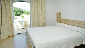 Quarto com cama, janela com cortinas, mesinha e vista para o verde lá fora.