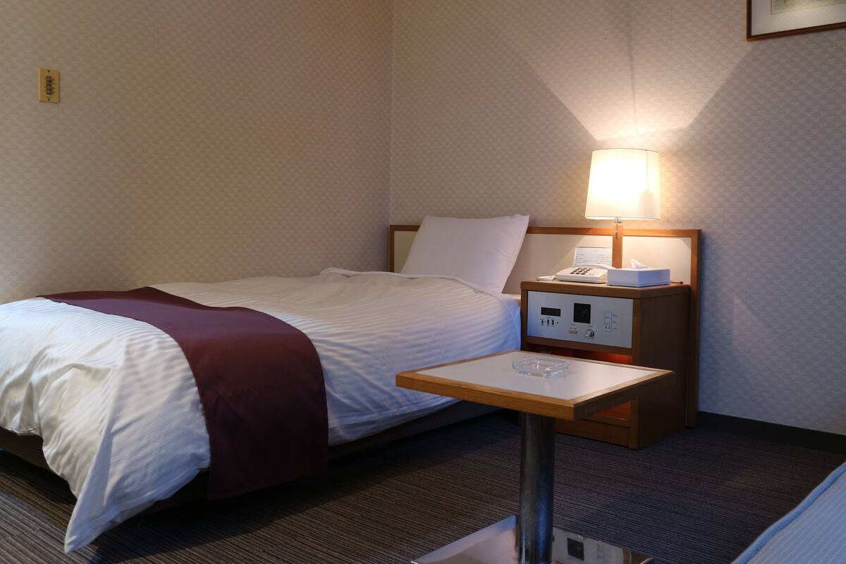 A hotel room with a bed, a bedside table with a lamp, and a small table with a glass.