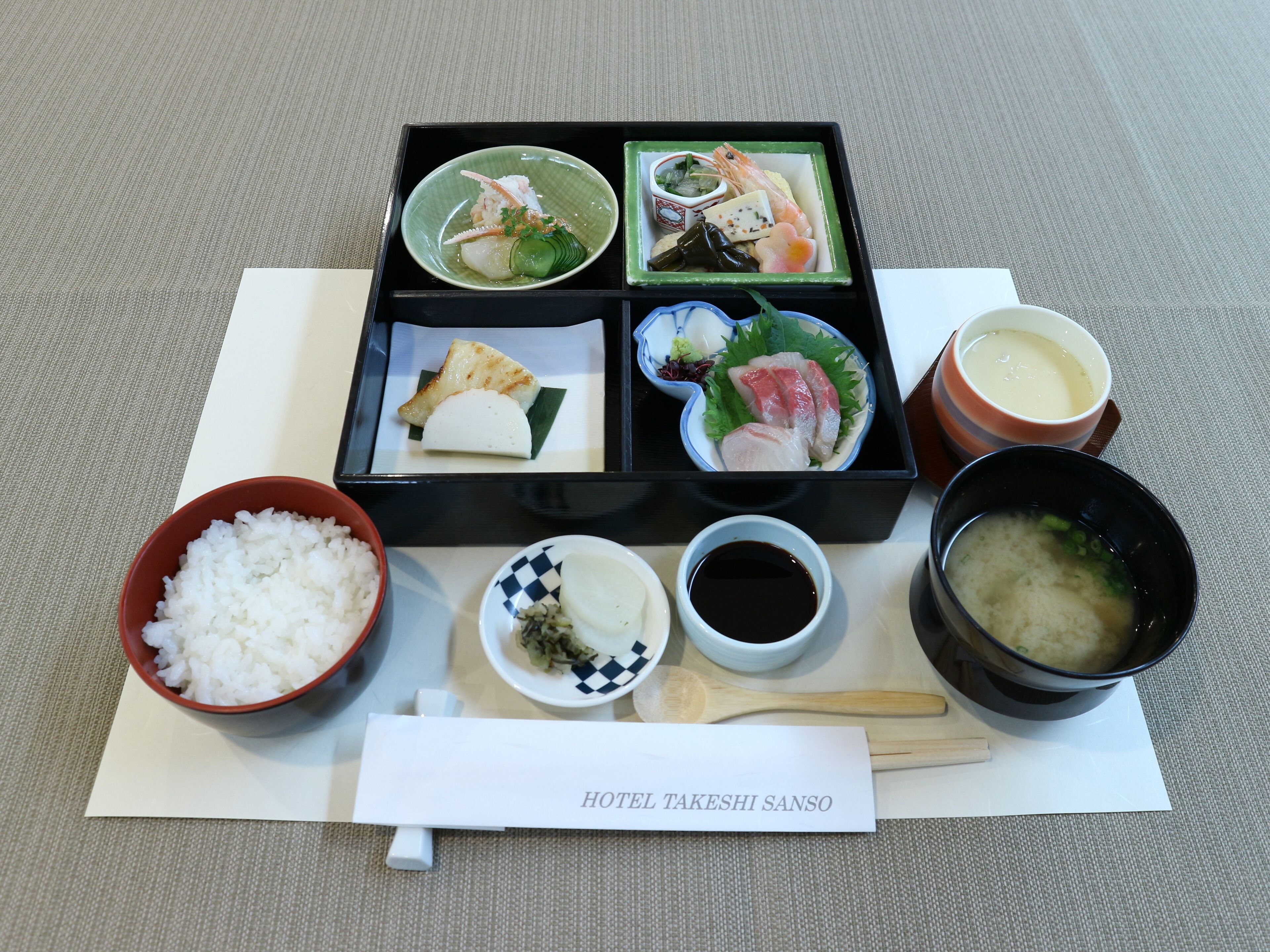 A traditional Japanese meal with rice, soup, and various dishes in a black bento box.