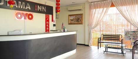 A hotel reception area with a reception desk, a bench, and a large window with sheer curtains.